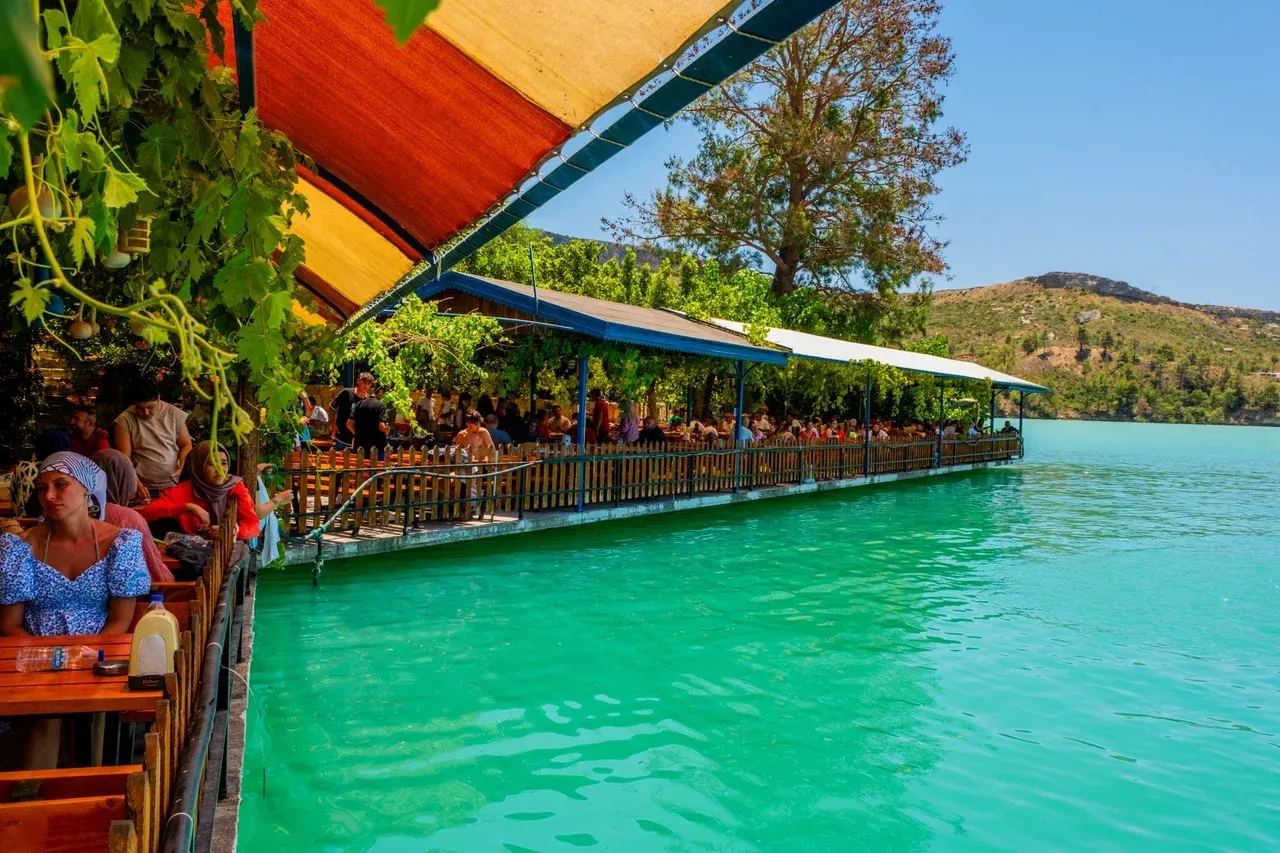 Restaurantterrasse unter orangefarbenen Markisen auf dem grünen Wasser des Yeşilgöl, dahinter kahle Hügel