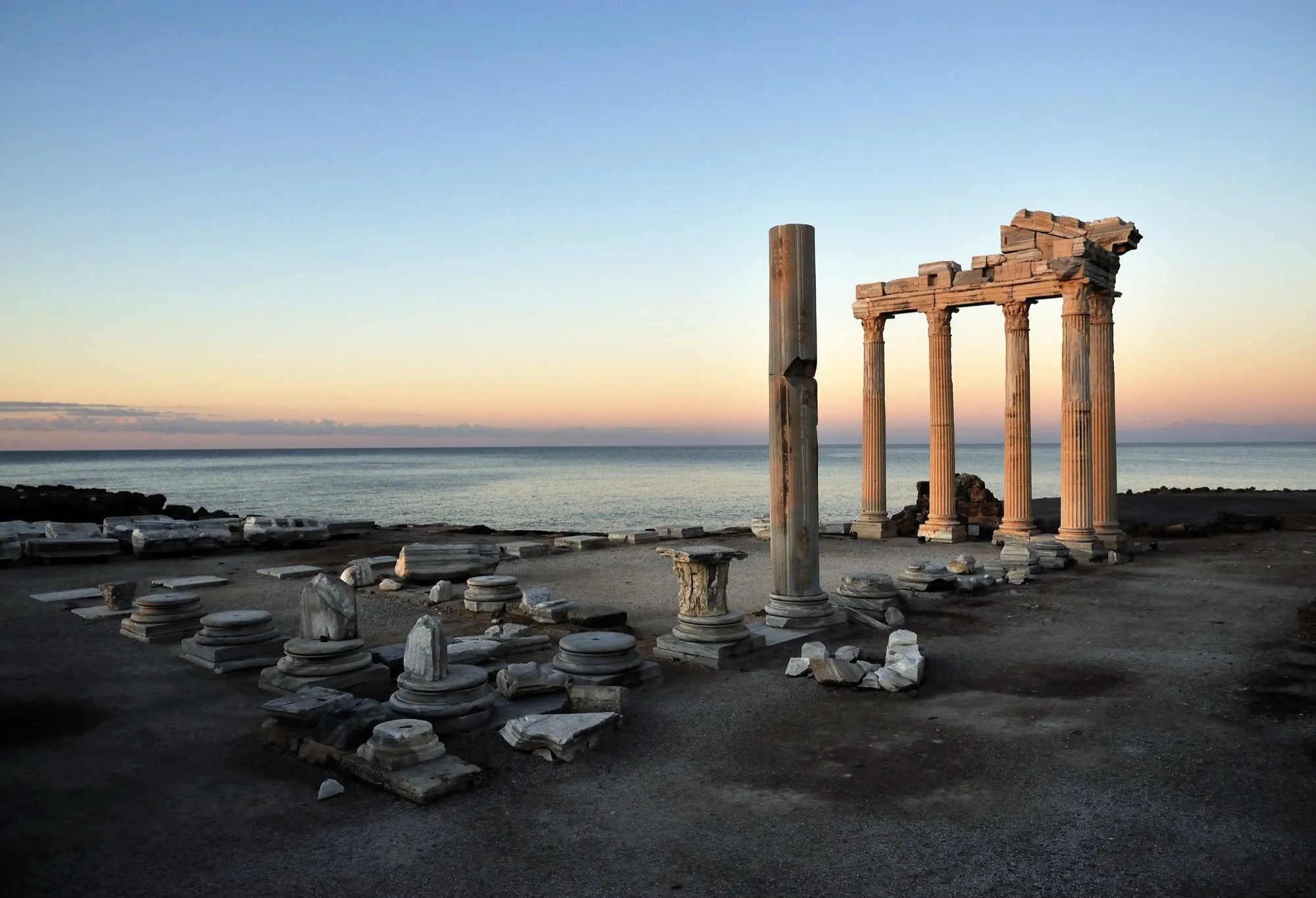 Der Apollon-Tempel in der Abenddämmerung: fünf beleuchtete Säulen vor pastellfarbenem Himmel, verstreute Säulenbasen und Kapitelle auf der dunklen Plattform, dahinter das ruhige Meer